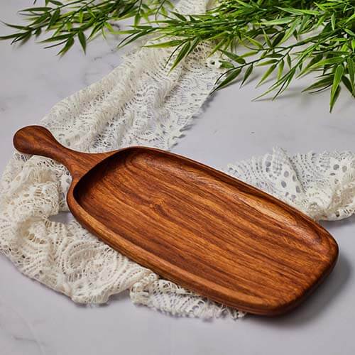 Handmade cheese serving tray made of rich, polished wood, placed on lace fabric with green leaves above.